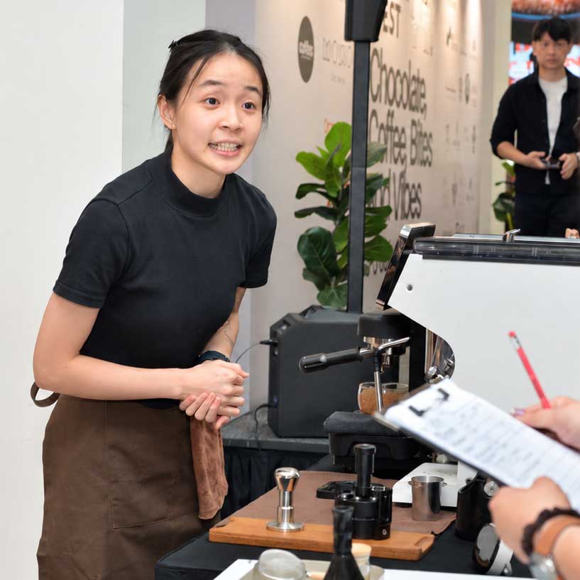 A confident barista contender, Josephine Yap Wei Yuen stands behind a coffee station with an espresso machine, during 2026 KL Mocha Fest competition. She ended up winning.